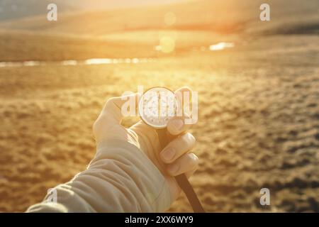 La main d'un homme tient une boussole à main sur fond de montagnes et de collines au coucher du soleil. Le concept de voyage et de navigation dans les espaces ouverts Banque D'Images