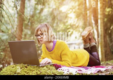Image tonifiée d'un portrait de fille freelance dans un pull jaune et des lunettes regardant soigneusement l'écran d'ordinateur portable dans la nature dans un front de conifères Banque D'Images