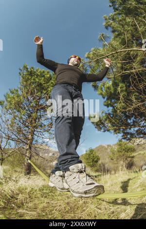 Un homme, vieilli avec une barbe et portant des lunettes de soleil, se balance sur une slackline en plein air entre deux arbres au coucher du soleil sur fond bleu ciel Banque D'Images