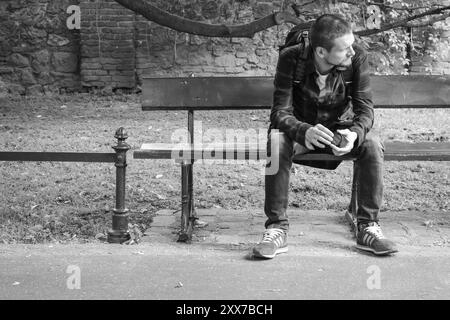 Homme solitaire assis sur le banc, monochrome. Touriste avec sac à dos et chaussures adidas pensant sur banc dans le parc, noir et blanc. Mode de vie actif. Banque D'Images