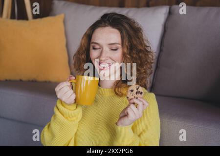 Photo de belle jeune femme se détendre manger collation boire du thé porter élégant pull jaune moderne lumière confortable intérieur plat à l'intérieur Banque D'Images