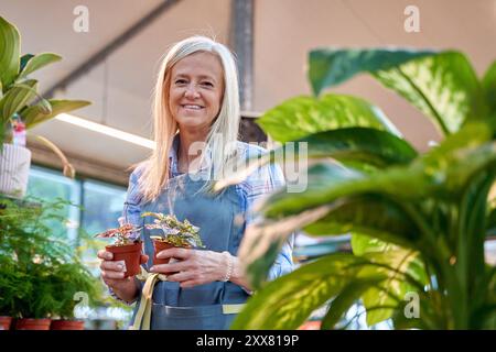 femme d'âge moyen travaillant dans un magasin de fleurs, regarde la caméra en souriant tout en plaçant deux petites plantes. Banque D'Images