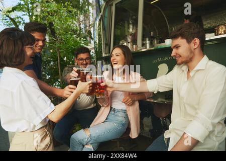 Groupe de jeunes heureux souriants, hommes et femmes, amis se réunissant au bar extérieur local, boire de la bière, clinking verres et avoir de bonnes vibrations de Banque D'Images