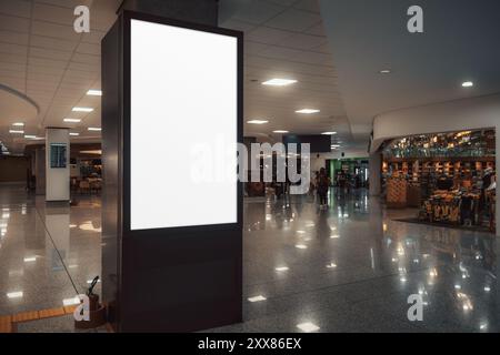 Une photo discrète d'une maquette de panneau d'affichage vide verticale se tient bien en vue au milieu d'un terminal d'aéroport animé ou d'un centre commercial, avec des magasins à l'intérieur Banque D'Images