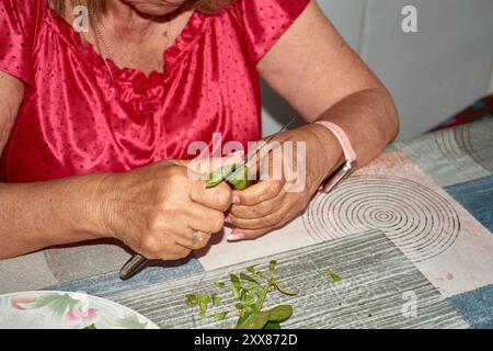 Les mains d'une femme âgée alors qu'elle tranche soigneusement les haricots verts avec un couteau, les préparant pour la cuisson. Les mains altérées mais habiles, marquées b Banque D'Images