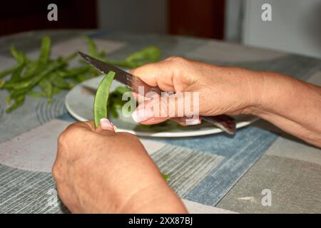 Les mains d'une femme âgée alors qu'elle tranche soigneusement les haricots verts avec un couteau, les préparant pour la cuisson. Les mains altérées mais habiles, marquées b Banque D'Images