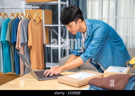 Heureux jeune homme vendeur en ligne vérifiant les commandes entrantes de son ordinateur portable à partir de son home studio Banque D'Images