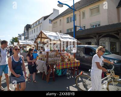 Hardelot, France - 20 août 2023 : des acheteurs et un cycliste passent devant des étals de marché à Neufchatel-Hardelot, France, où un vendeur expose une variété de miel et d'autres produits sous le soleil d'été Banque D'Images