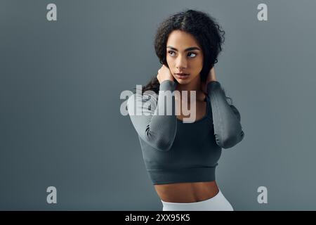 Une jeune femme dans un haut gris se tient devant un fond gris, regardant sur le côté et ajustant ses cheveux. Banque D'Images