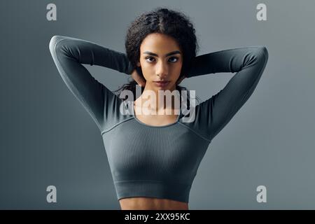 Une jeune femme afro-américaine aux cheveux bouclés étire ses bras au-dessus de la tête, portant un haut gris à manches longues sur un fond gris. Banque D'Images