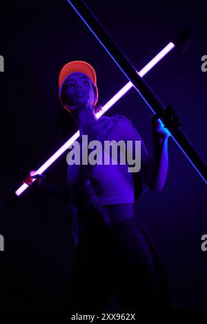 Une jeune femme dans un studio, baignée de néons violets, tient une lampe brillante dans une pose confiante et élégante. Banque D'Images