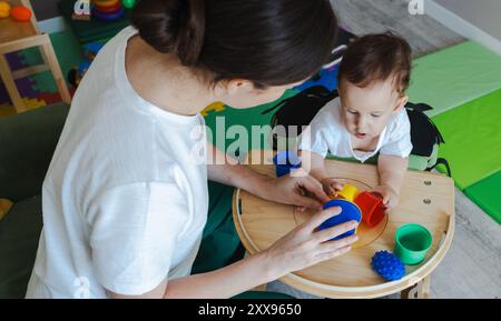 Le patient petit enfant masculin s'engage dans une session de réadaptation physique avec un physiothérapeute, interagissant avec des jouets colorés sur la table de jeu Banque D'Images