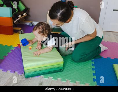 Garçon avec faiblesse musculaire portant corset colonne vertébrale s'engage dans des exercices de réadaptation avec un thérapeute, en utilisant des tapis colorés et des équipements de soutien Banque D'Images