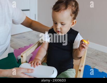 Mignon petit garçon portant corset de réadaptation s'engage dans la thérapie physique, se concentrant sur des exercices de mobilité tout en étant assis dans une chaise de soutien avec un th physique Banque D'Images