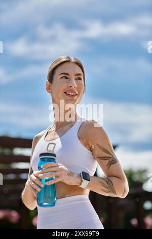 Une jeune femme en tenue d'action, avec du vitiligo, sourit et tient une bouteille d'eau tout en s'entraînant dans un parc. Banque D'Images