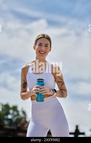 Une jeune femme en vêtements d'activité blancs sourit et tient une bouteille d'eau tout en prenant une pause de son entraînement en plein air. Banque D'Images