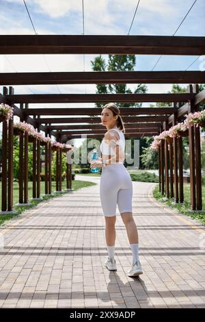 Une jeune sportive, vêtue de vêtements de sport blancs, marche le long d'un chemin de briques sous une pergola en bois, tenant une bouteille d'eau. Banque D'Images