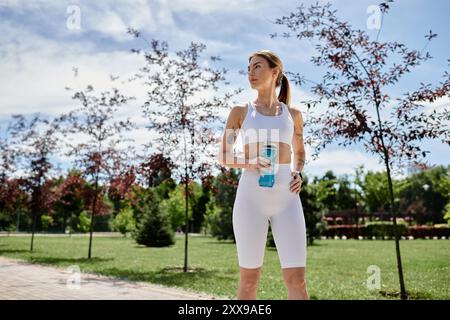 Une jeune femme atteinte de vitiligo, vêtue de vêtements de sport blancs, interrompt son entraînement dans un parc, tenant une bouteille d'eau. Banque D'Images