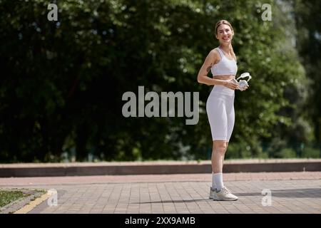 Une sportive souriante en vêtements actifs blancs tient ses écouteurs, prenant une pause de son entraînement dans un parc Banque D'Images