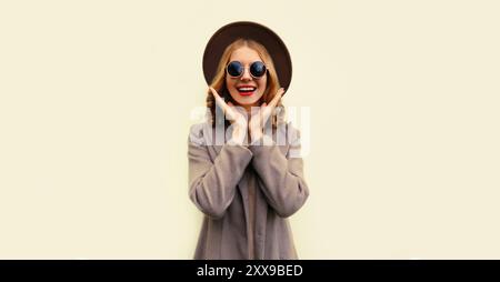 Portrait d'un beau modèle jeune femme souriant portant un chapeau rond brun et un manteau sur fond beige Banque D'Images