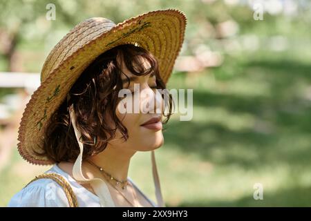 Une jeune femme aux cheveux bruns dans un chapeau de paille et un chemisier blanc se détend dans un jardin ensoleillé, les yeux fermés dans une contemplation paisible. Banque D'Images