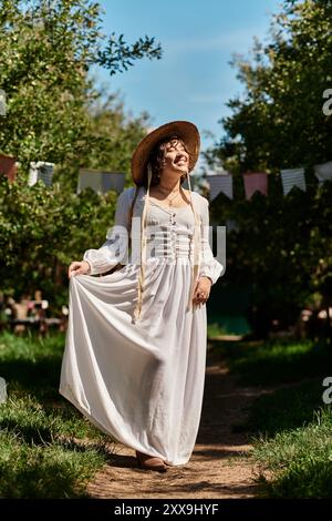 Une jeune femme vêtue d'une robe blanche et d'un chapeau de paille marche le long d'un chemin dans un jardin, entourée d'arbres verts et d'un ciel bleu. Banque D'Images