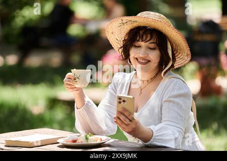 Une jeune femme dans une robe blanche et un chapeau de paille profite d'un brunch tranquille dans un jardin d'été, regardant le téléphone. Banque D'Images