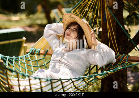 Une jeune femme en robe blanche et chapeau de paille se détend dans un hamac sous un ciel d'été. Banque D'Images