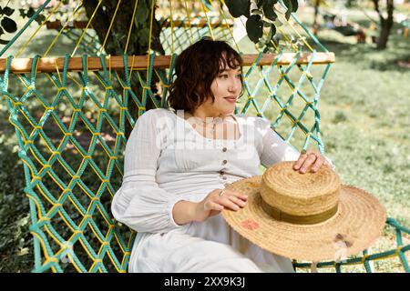 Une jeune femme en robe blanche et chapeau de paille se détend dans un hamac à l'ombre d'un arbre. Banque D'Images