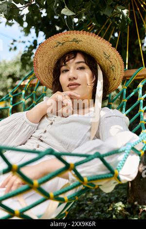 Une jeune femme en robe blanche et chapeau de paille se détend dans un hamac entouré de verdure. Banque D'Images