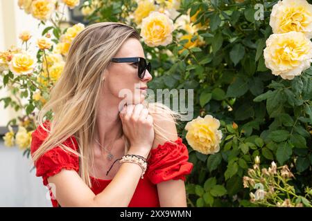 Jeune femme blonde caucasienne en lunettes de soleil entourée de roses jaunes dans un jardin Banque D'Images