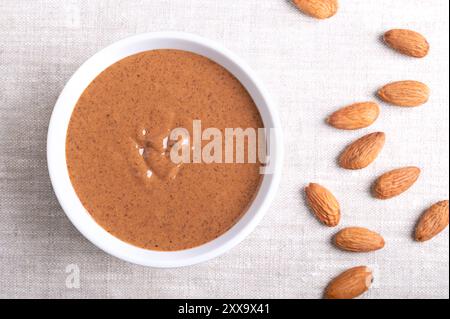 Beurre d'amande dans un bol blanc sur tissu de lin. Pâte alimentaire brune et lisse, fabriquée à partir du broyage d'amandes non écaillées en beurre de noix. Banque D'Images