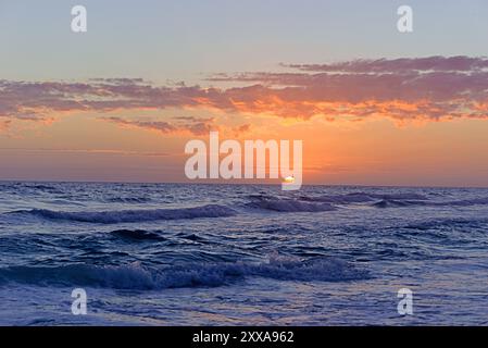 Un beau coucher de soleil projette une lueur orange et rose sur les vagues de l'océan, avec le soleil apparaissant à mi-chemin sous l'horizon. Banque D'Images