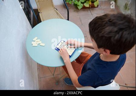 Un jeune garçon joue aux dominos sur une table bleue à l'extérieur. La scène représente un moment calme et engageant, parfait pour les amateurs de loisirs et de stratégie. Banque D'Images