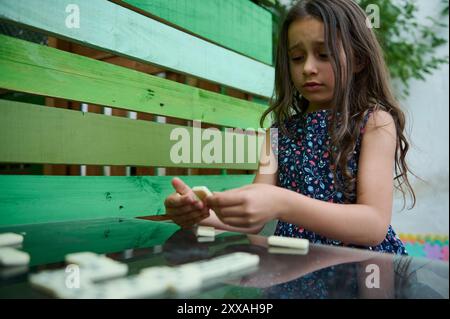 Une jeune fille dans une robe fleurie joue des dominos à l'extérieur, profondément absorbée par la pensée alors qu'elle envisage son prochain mouvement stratégique. Banque D'Images