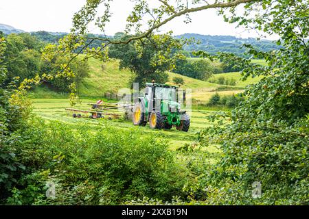 Tournage de l'herbe pour l'ensilage avec un tracteur John Deere 6130R et un râteau à foin Claas Liner 2900 à Irthington, Cumbria, Angleterre, Royaume-Uni Banque D'Images