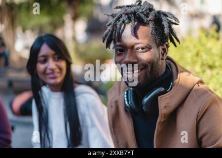 Portrait d'un homme africain joyeux avec des dreadlocks portant des écouteurs, accompagné d'une femme souriante aux longs cheveux foncés. Couple diversifié appréciant urb Banque D'Images