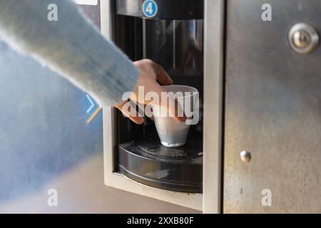 Gros plan de la main d'une personne cherchant un gobelet jetable rempli de café à partir d'un distributeur automatique. Confort moderne sur le lieu de travail ou dans un pub Banque D'Images