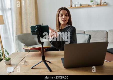Une jeune femme travaille à distance, filmant une vidéo sur son téléphone tout en étant assise à son bureau avec un ordinateur portable. Banque D'Images