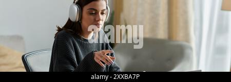 Une jeune femme portant des écouteurs travaille sur un ordinateur portable à son bureau à domicile, avec une expression ciblée. Banque D'Images