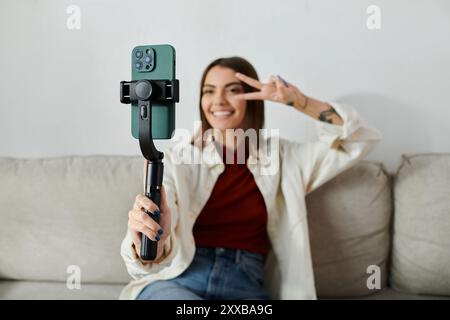 Une jeune femme vêtue de façon décontractée filme du contenu pour son vlog en utilisant un stabilisateur de smartphone, assise sur un canapé. Banque D'Images