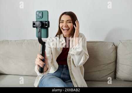 Une jeune femme au sourire éclatant est assise sur un canapé, filmant un vlog avec son smartphone et son stabilisateur. Banque D'Images