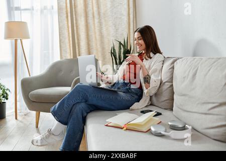 Une jeune femme sourit alors qu'elle travaille sur son ordinateur portable depuis le canapé de son salon, écouteurs à proximité. Banque D'Images