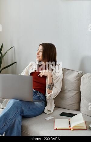 Une jeune femme en tenue décontractée est assise sur un canapé avec un ordinateur portable sur ses genoux, perdue dans ses pensées de sa prochaine campagne sur les médias sociaux. Banque D'Images