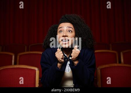 Une jeune femme est assise dans un siège de théâtre, ses mains serrées devant elle alors qu'elle réagit à un film qui passe à l'écran. Banque D'Images