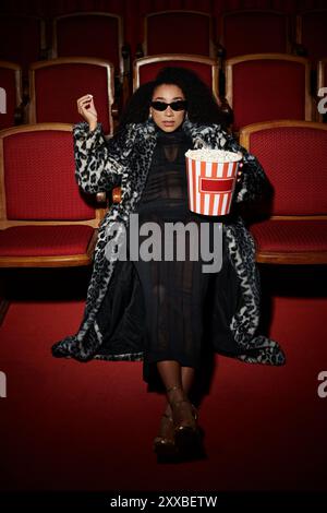 Une femme afro-américaine élégante portant un manteau imprimé léopard regarde un film dans un cinéma sombre. Banque D'Images