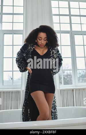 Une femme afro-américaine élégante dans une robe noire et un manteau léopard pose dans un cadre de studio, créant une image chic et moderne. Banque D'Images