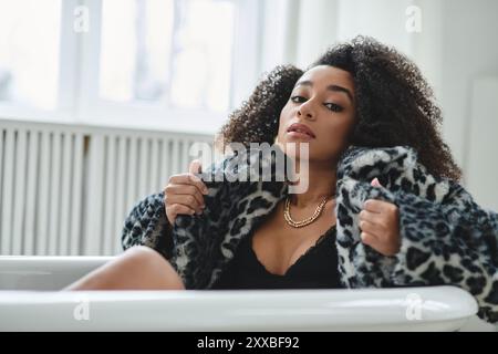 Une femme avec un afro dans un manteau à imprimé léopard à la mode est assise dans une baignoire blanche et regarde directement la caméra. Banque D'Images