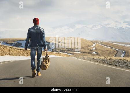 Un hipster barbu élégant dans des lunettes de soleil avec un sac à dos vintage marche le long de la route asphaltée par une journée ensoleillée. Le concept d'auto-stop et de randonnée Banque D'Images