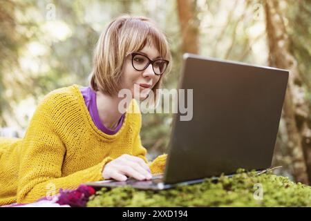 Image tonifiée d'un portrait de fille freelance dans un pull jaune et des lunettes regardant soigneusement l'écran d'ordinateur portable dans la nature dans un front de conifères Banque D'Images